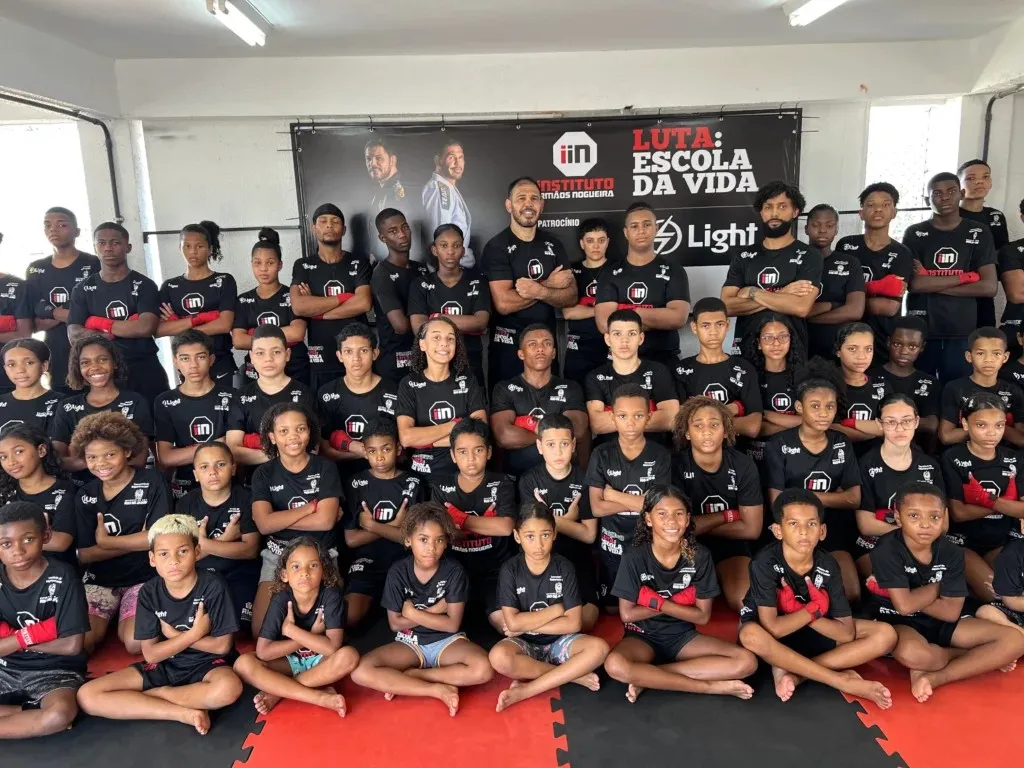 Turma do Luta Escola da Vida reunida no tatame, de camiseta do Instituto e luvas de Muay Thai, diante do banner do projeto